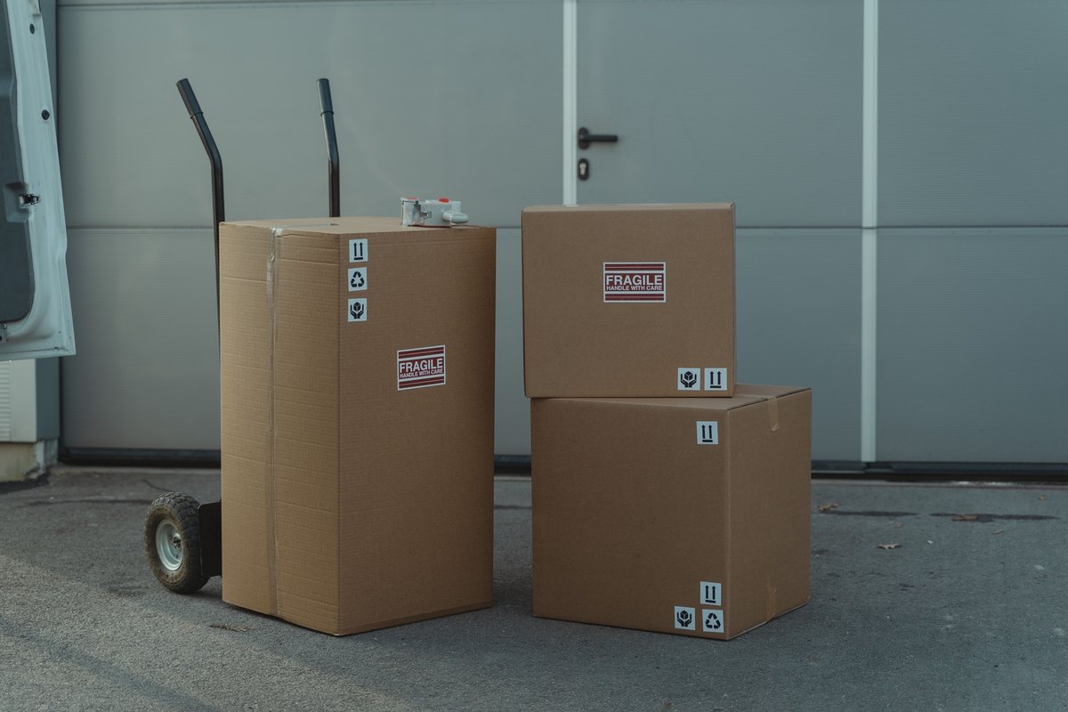 Stacked cardboard boxes with fragile labels and a hand truck on pavement in front of a gray garage door