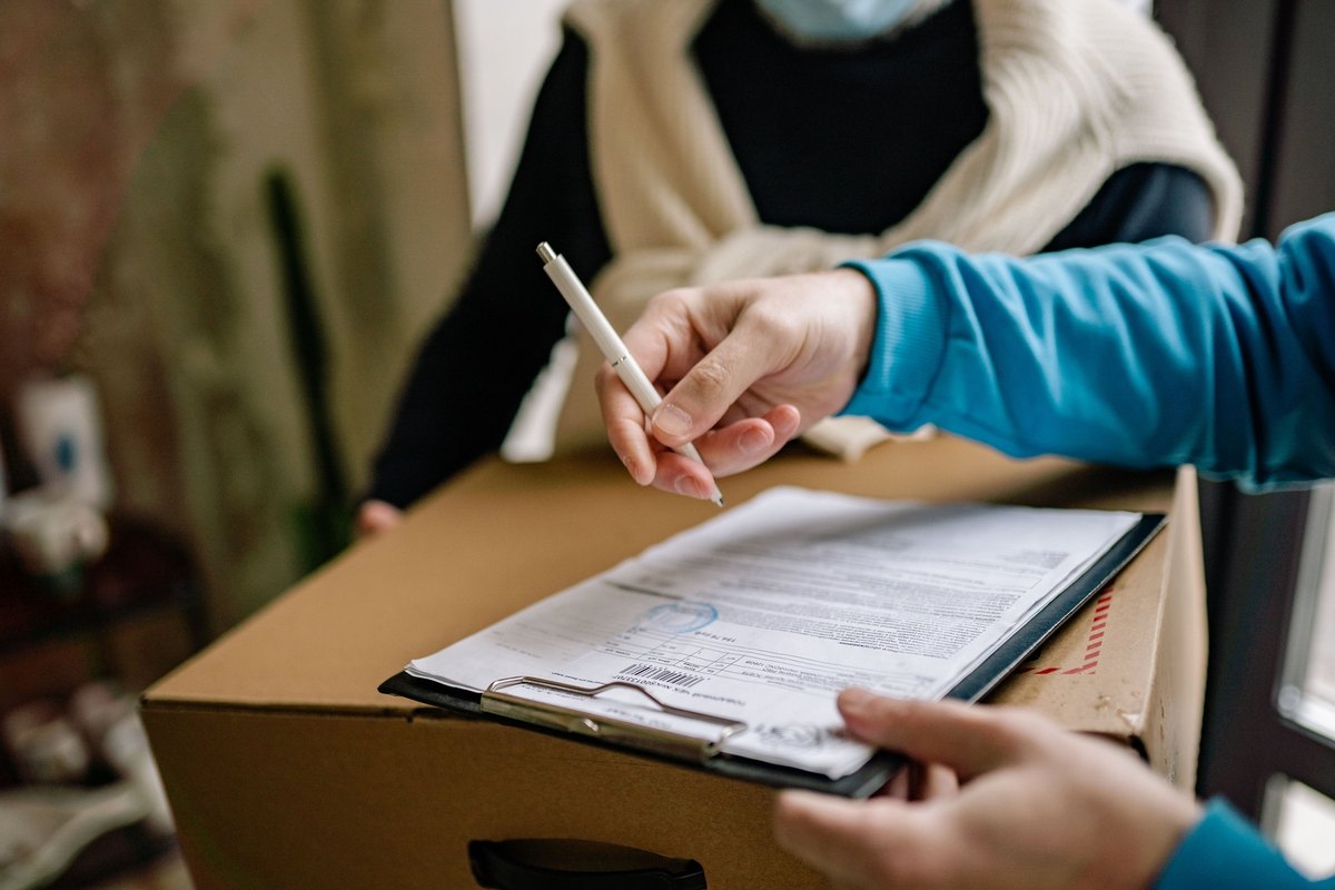 Hand signing a delivery form on a clipboard resting on a cardboard box