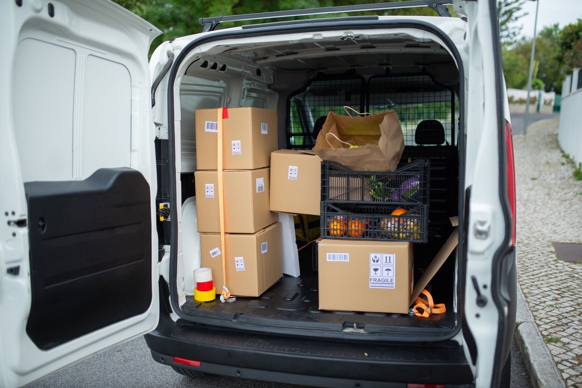 Open white delivery van loaded with stacked cardboard boxes and a crate of produce