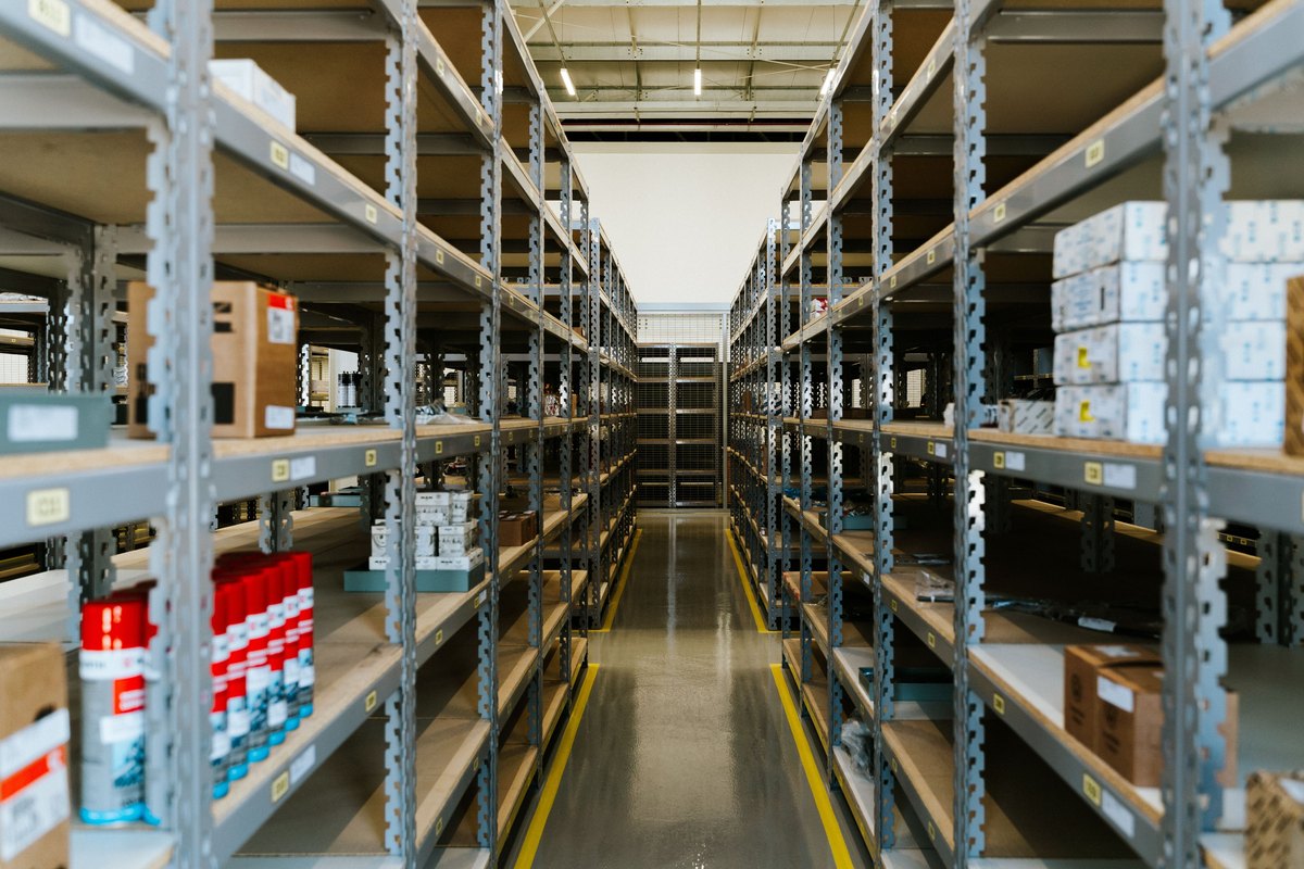 Long aisle between gray metal shelving holding cartons and parts boxes in a bright warehouse